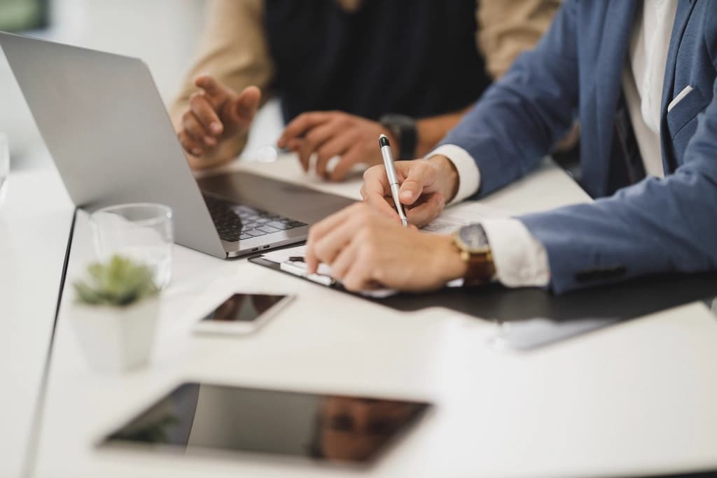 Two colleagues at a white desk reviewing documents beside laptops